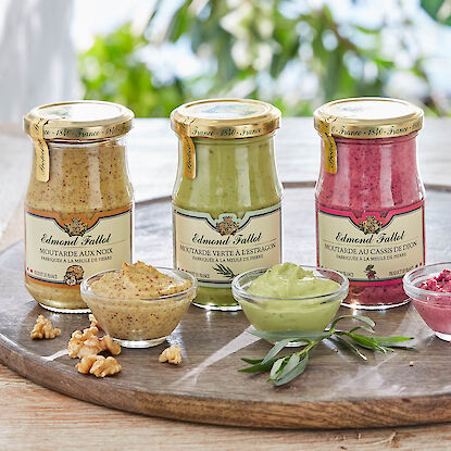 Trois pots de moutarde Edmond Fallot aux noix, à l’estragon et au cassis avec des coupelles de moutarde sur une planche en bois.