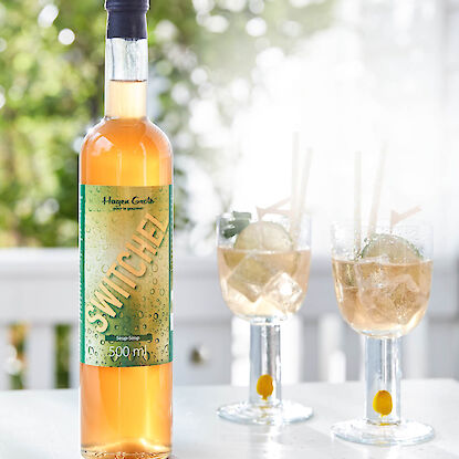 Bouteille de sirop Switchel Hagen Grote avec deux verres de boisson estivale, glaçons, rondelles de citron vert et menthe sur table blanche.