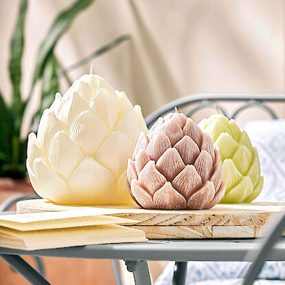 Drei Kerzen in Artischockenform in Creme, Rosa und Grün auf Holzbrett auf Gartentisch.