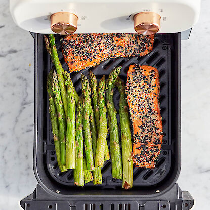 Filets de saumon en croûte de sésame et asperges vertes dans une friteuse à air chaud sur plan de travail en marbre.Schwarze Heißluftfritteuse mit zwei Fächern, befüllt mit Gemüse und Kartoffel-Waffelpommes, daneben servierte Gerichte.