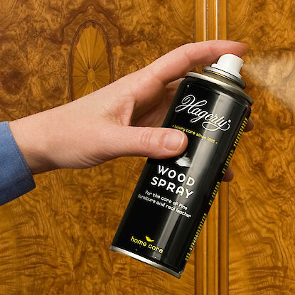 Eine Hand hält eine schwarze Sprühdose mit der Aufschrift „Hagerty Wood Spray“ vor eine glänzende, gemaserte Holzoberfläche. Aus der Dose wird ein feiner Sprühnebel abgegeben. Die Dose ist beschriftet mit dem Text „For the care of fine furniture and real leather“. Im Hintergrund ist eine edel furnierte Holztür oder ein Möbelstück mit auffälliger Maserung und Intarsien zu sehen.