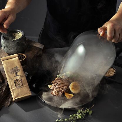 Un chef soulève un dôme en verre rempli de fumée au-dessus d’une assiette de viande grillée, d’oignons et d’herbes. À côté se trouvent un mortier avec des épices, un couteau, de l’ail rôti, du sel, du poivre et un sac en papier brun portant l’inscription « Olive – Natural 100% Quality ». La scène présente une élégante mise en valeur culinaire avec un effet de fumée.