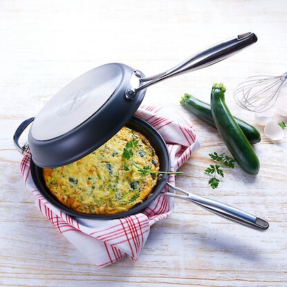 Une frittata aux légumes sautés dans une poêle noire avec un couvercle en acier inoxydable, présentée sur un torchon de cuisine à carreaux rouges et blancs. À côté, des courgettes fraîches, des coquilles d'œufs, un fouet et du persil reposent sur une surface en bois blanc – des ingrédients qui évoquent une préparation maison et fraîche.