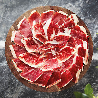 Jambon ibérique espagnol finement tranché, joliment disposé sur une planche en bois ronde, avec un fond en pierre naturelle sombre.