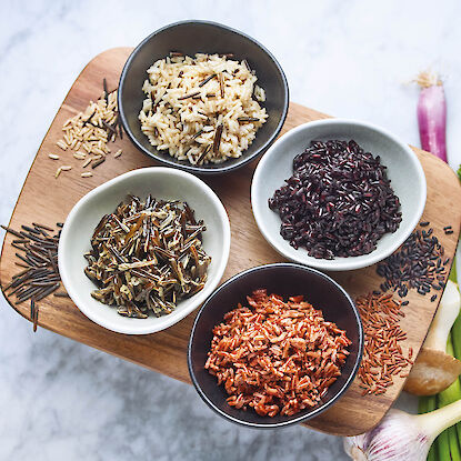 Quatre variétés de riz – riz sauvage, riz noir, riz rouge et mélange de riz sauvage – joliment présentées dans des petits bols sur une planche en bois, accompagnées d’ingrédients crus comme l’ail et les oignons nouveaux.