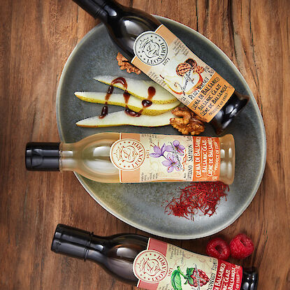 Trois bouteilles de crème de balsamique Leonardi aux saveurs poire-noix, safran et framboise sur une assiette grise avec des tranches de poire, des noix, des filaments de safran et des framboises fraîches, le tout posé sur une table en bois.