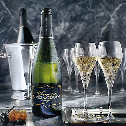 Une élégante bouteille de champagne avec de la condensation est posée sur une surface en marbre. À côté se trouvent deux flûtes à champagne remplies sur un plateau en argent. En arrière-plan, un seau à champagne avec une autre bouteille ainsi que quelques verres vides sont visibles. Un bouchon de champagne repose sur la table à l'avant.