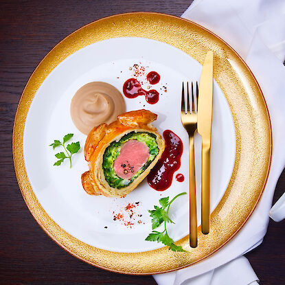 Plat élégant sur une assiette blanche à bord doré, composé d'un morceau de filet Wellington avec des légumes verts, une sauce crémeuse brune, une touche de sauce aux fruits rouges et des herbes fraîches. Des couverts en métal doré sont placés à droite du plat sur une serviette en tissu blanche sur une table en bois foncé.