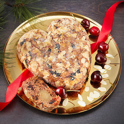 Stollen en forme de cœur avec cerises et pâte d'amande sur une assiette dorée, décorée d'un ruban rouge.