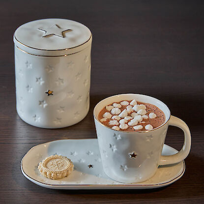 Weiße Tasse und Vorratsdose mit Sternmotiv, gefüllt mit Kakao und Marshmallows, auf Teller mit Gebäck