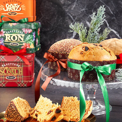 Panettone mit bunten Schleifen auf Glasetagere, daneben festliche Geschenkboxen und Zweige