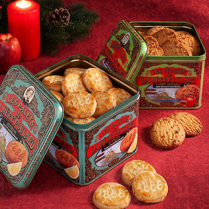 Bunte Metalldosen mit Buttergebäck auf rotem Tischtuch, Kerze und Apfel im Hintergrund