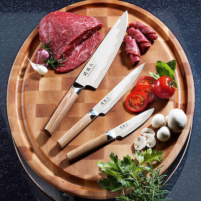 Drei japanische Kochmesser mit Fleisch, Tomaten und Pilzen auf Holzbrett arrangiert