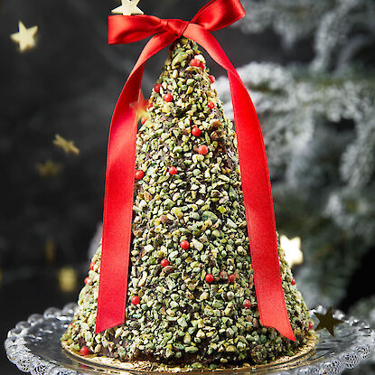 Weihnachtstorte in Tannenbaumform mit Pistazien und roter Schleife auf Glasplatte.