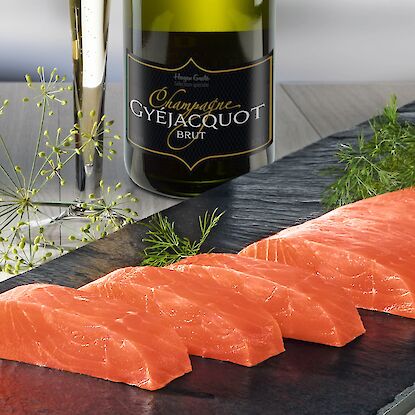 Lachsscheiben auf Schieferplatte mit Dill, dazu Champagnerflasche und gefülltes Glas im Hintergrund.