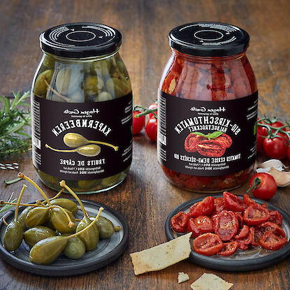 Zwei Gläser mit Kapernbeeren und halbgetrockneten Kirschtomaten, angerichtet auf kleinen Tellern mit Snacks.