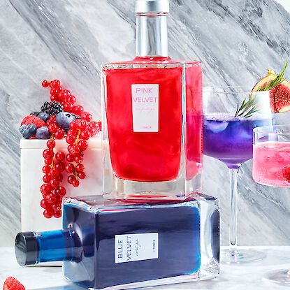 Pink- und Blue-Velvet-Gin in eckigen Flaschen, dekoriert mit Beeren und farbigen Drinks im Hintergrund.