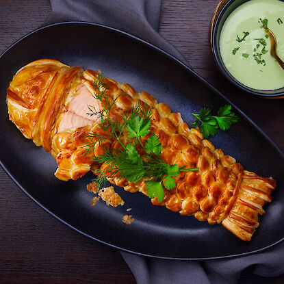 Lachs im Blätterteig in Fischform gebacken – serviert mit Kräutern und grüner Sauce auf dunklem Teller