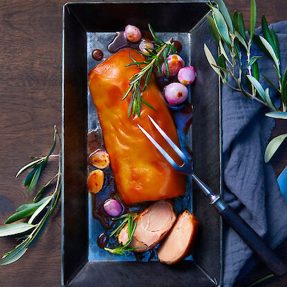 Saftiger Braten mit goldbrauner Kruste, Schalotten und Rosmarin in Metallform, angerichtet auf dunklem Holztisch