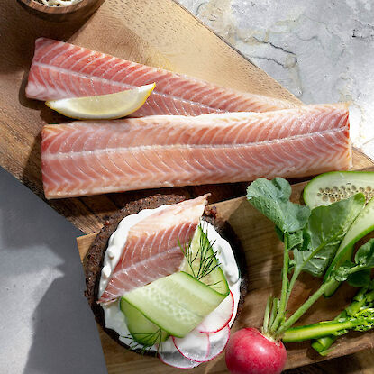 Zarte, geräucherte Fischfilets mit Zitrone auf Holzbrett, dazu Brot mit Creme, Gurke, Radieschen und frischen Kräutern