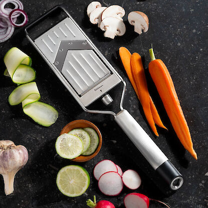 Gemüsereibe aus Metall mit geschnittenen Karotten, Zucchini, Radieschen, Pilzen, Zwiebeln, Knoblauch und Limetten auf dunkler Fläche