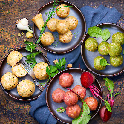 Verschiedene Knödelvarianten auf Tellern – Natur, Spinat und Rote Bete – dekoriert mit Kräutern, Knoblauch, Parmesan und Gemüse