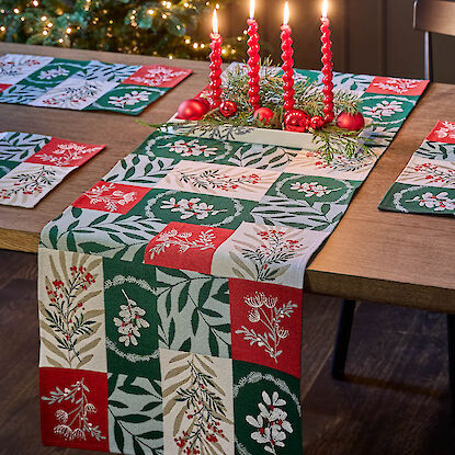 Weihnachtlicher Tischläufer in Rot, Grün und Creme mit Pflanzenmotiven, dekoriert mit roten Kerzen und Tannenzweigen auf einem Holztisch.