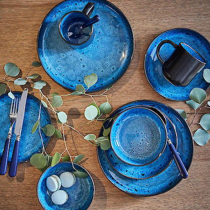 Blaues Keramikgeschirr im Set mit Tellern, Schalen und Tassen auf Holztisch, dekoriert mit Eukalyptuszweigen und Macarons.