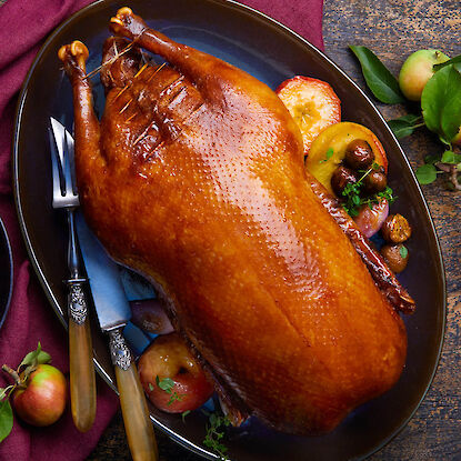 Gebratene ganze Ente auf Servierplatte mit Bratäpfeln und Esskastanien, angerichtet mit Besteck und herbstlicher Deko.