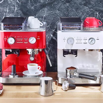Zwei KitchenAid-Siebträgermaschinen in Rot und Weiß auf Arbeitsplatte, umgeben von Kaffeezubehör und Bohnen.