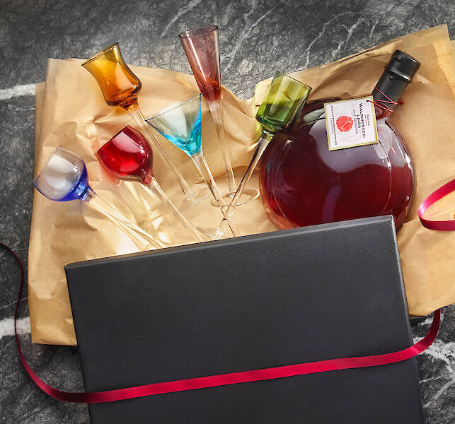 Geschenkset mit farbigen Likörgläsern und einer bauchigen Flasche Likör, arrangiert auf Papier mit schwarzer Geschenkbox.