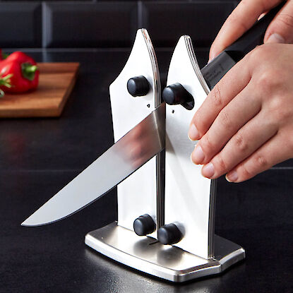 Nutzer schärft ein Küchenmesser in einem manuellen Messerschärfer aus Metall auf dunkler Arbeitsfläche.