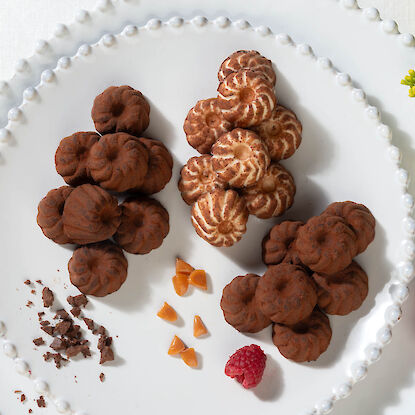 Pralinen in Gugelhupf-Form auf weißem Teller, dekoriert mit Schokostückchen, Karamell und Himbeere.