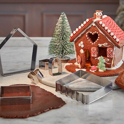 Ausstechformen und Lebkuchenhaus auf Marmorplatte, mit Teig, Deko und kleinem Tannenbaum.