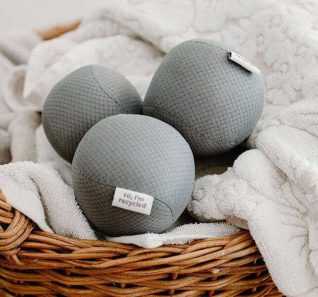 Drei graue Trocknerb&auml;lle aus recyceltem Material in einem W&auml;schekorb mit weichen Handt&uuml;chern.