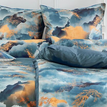 Bett mit blau-goldener Bettw&auml;sche im Wolkenmuster, dekoriert mit Kissen, Vasen und Nachttischen.