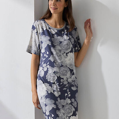 Frau tr&auml;gt ein knielanges Kleid mit blau-wei&szlig;em Blumenmuster und steht l&auml;chelnd an einer Wand.