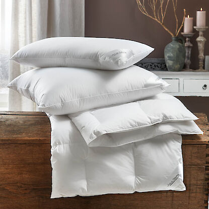 Wei&szlig;e Kopfkissen und Bettdecke auf einer Holztruhe, dekoriert mit Kerzen und Vase im Hintergrund.