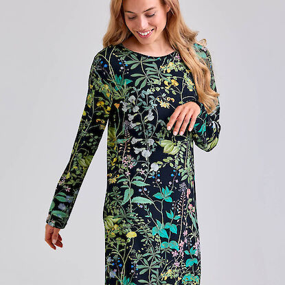 Frau tr&auml;gt ein schwarzes Kleid mit buntem botanischem Blumen- und Bl&auml;ttermuster und l&auml;chelt.