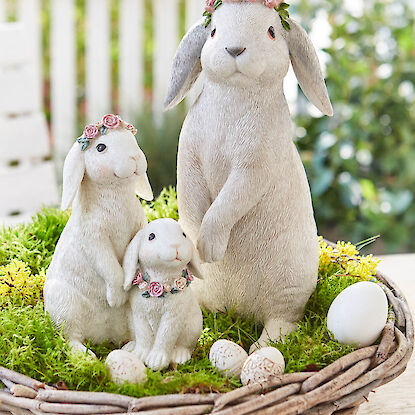Drei Hasenfiguren mit Blumenkr&auml;nzen in einem Nest aus Moos und Zweigen, dekoriert mit Eiern.
