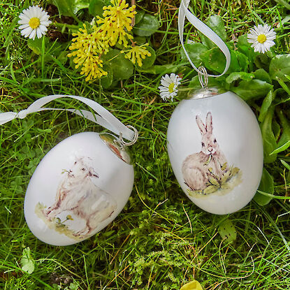 Zwei bemalte Ostereier mit Lamm- und Hasenmotiv, liegend im Gras mit Blumen und wei&szlig;en B&auml;ndern.