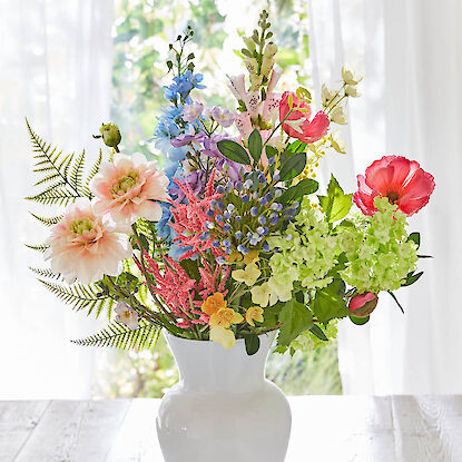 Bunter Fr&uuml;hlingsstrau&szlig; mit verschiedenen Blumen in einer wei&szlig;en Vase auf hellem Tisch vor Fenstern.