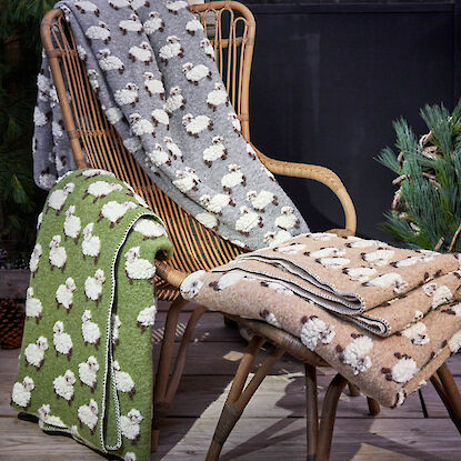 Wollige Decken mit Schafmotiven in Grau, Gr&uuml;n und Beige auf Rattanstuhl und Hocker arrangiert.