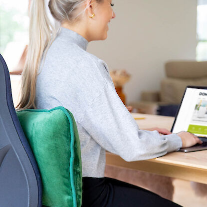 Frau sitzt am Schreibtisch und nutzt ein gr&uuml;nes Lendenkissen zur R&uuml;ckenst&uuml;tze am B&uuml;rostuhl.