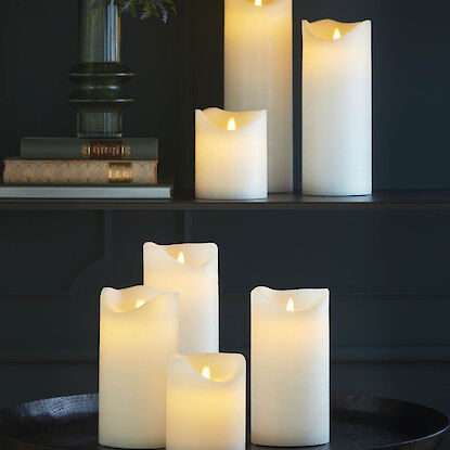 Mehrere LED-Kerzen in verschiedenen Gr&ouml;&szlig;en erzeugen warmes Licht auf Tisch und Regal.