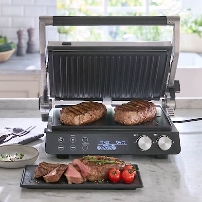 Gril de contact dans une cuisine moderne avec deux steaks grill&eacute;s sur la plaque, devant steak tranch&eacute; avec tomates.