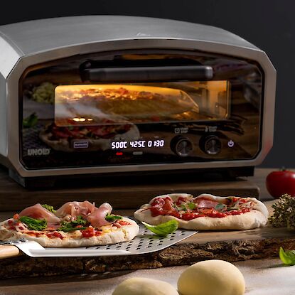 Four &agrave; pizza avec chambre visible, devant pizzas fra&icirc;chement garnies sur pelle, avec tomates et p&acirc;tons en pr&eacute;paration.