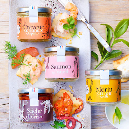 Quatre bocaux de tartinades marines sur un plateau, avec crevette, saumon, seiche, merlu, herbes fra&icirc;ches et crackers.