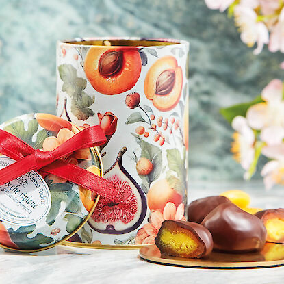 Dekorative Geschenkdose mit Fruchtmotiv und Schokot&ouml;rtchen, eines aufgeschnitten, auf Marmorplatte mit Blumen im Hintergrund.