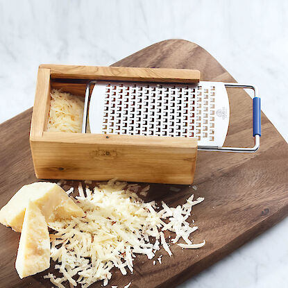 R&acirc;pe &agrave; fromage en bois avec grille, pos&eacute;e sur planche, avec fromage dur fra&icirc;chement r&acirc;p&eacute; et &eacute;clats sur surface marbr&eacute;e.